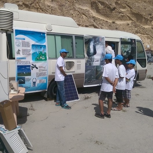Students listen to a teacher while standing near a RV with science posters affixed to it.