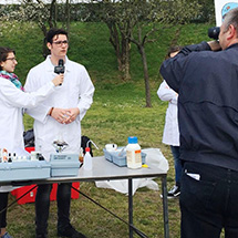 A man in a lab coat is interviewed by a woman with a microphone while a man with a camera records the interaction.