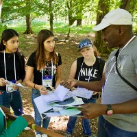 Three girl students are being taught by a man with books in his hand outside.
