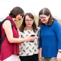 Three women are outside, standing and looking at a phone. One is pointing at the phone.