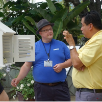 Two men are talking outside, with large leaves behind them. They are talking in front of a small white box that has an open door.