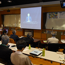 A group watches a large screen displaying a video address.