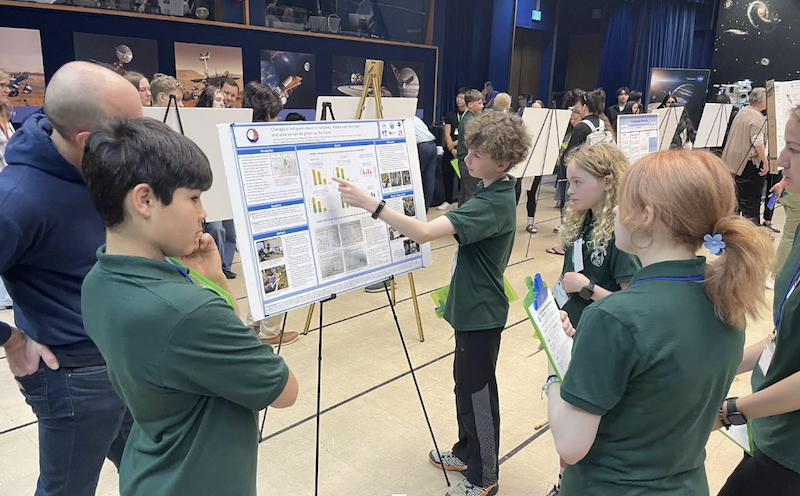 students stand in a room full of research posters on easels to present their local research