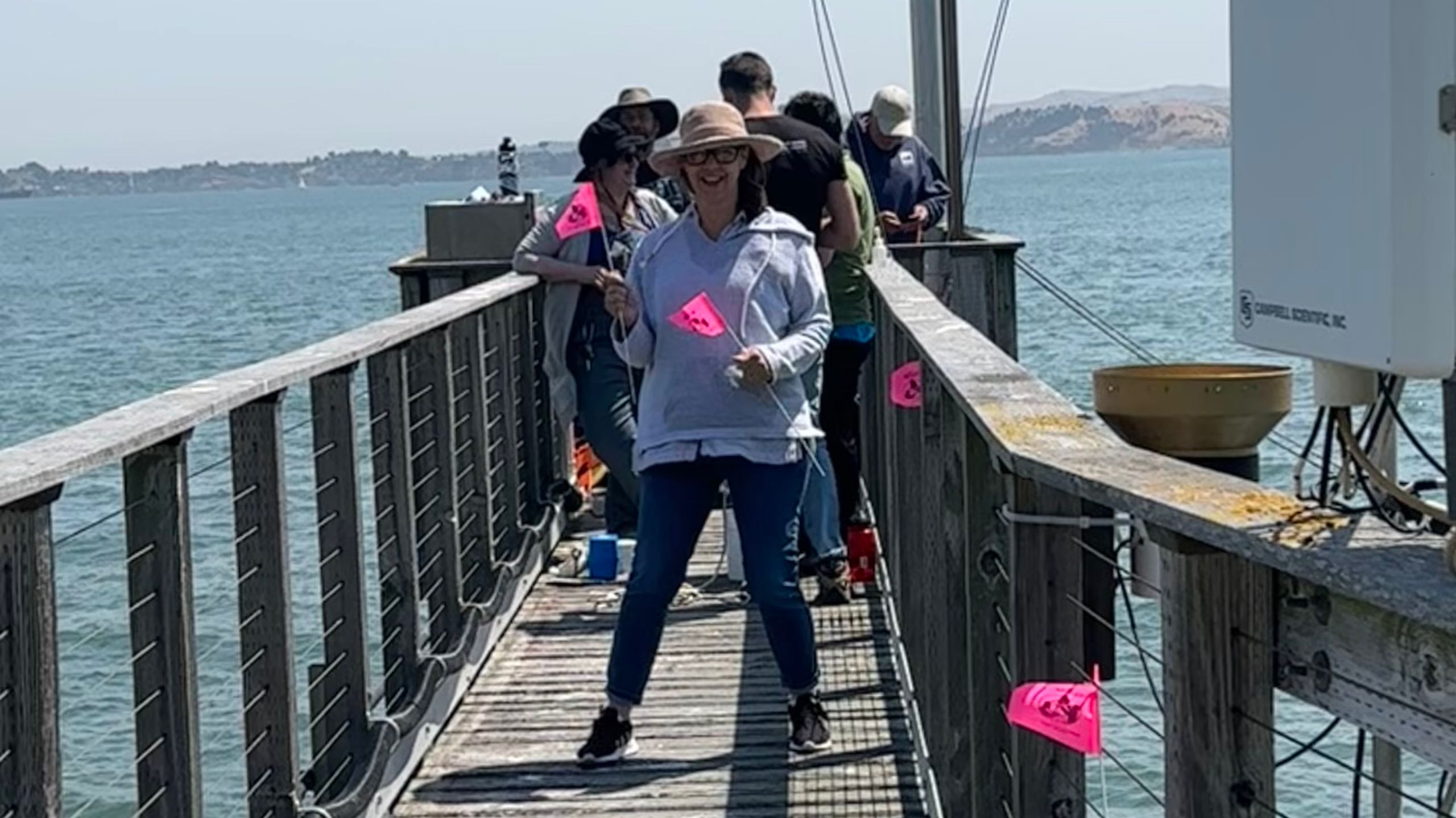 a woman stands on a pier, facing the camera, smiling and holding pink flags with the GLOBE logo while a group of educators stands behind her taking measurements of water