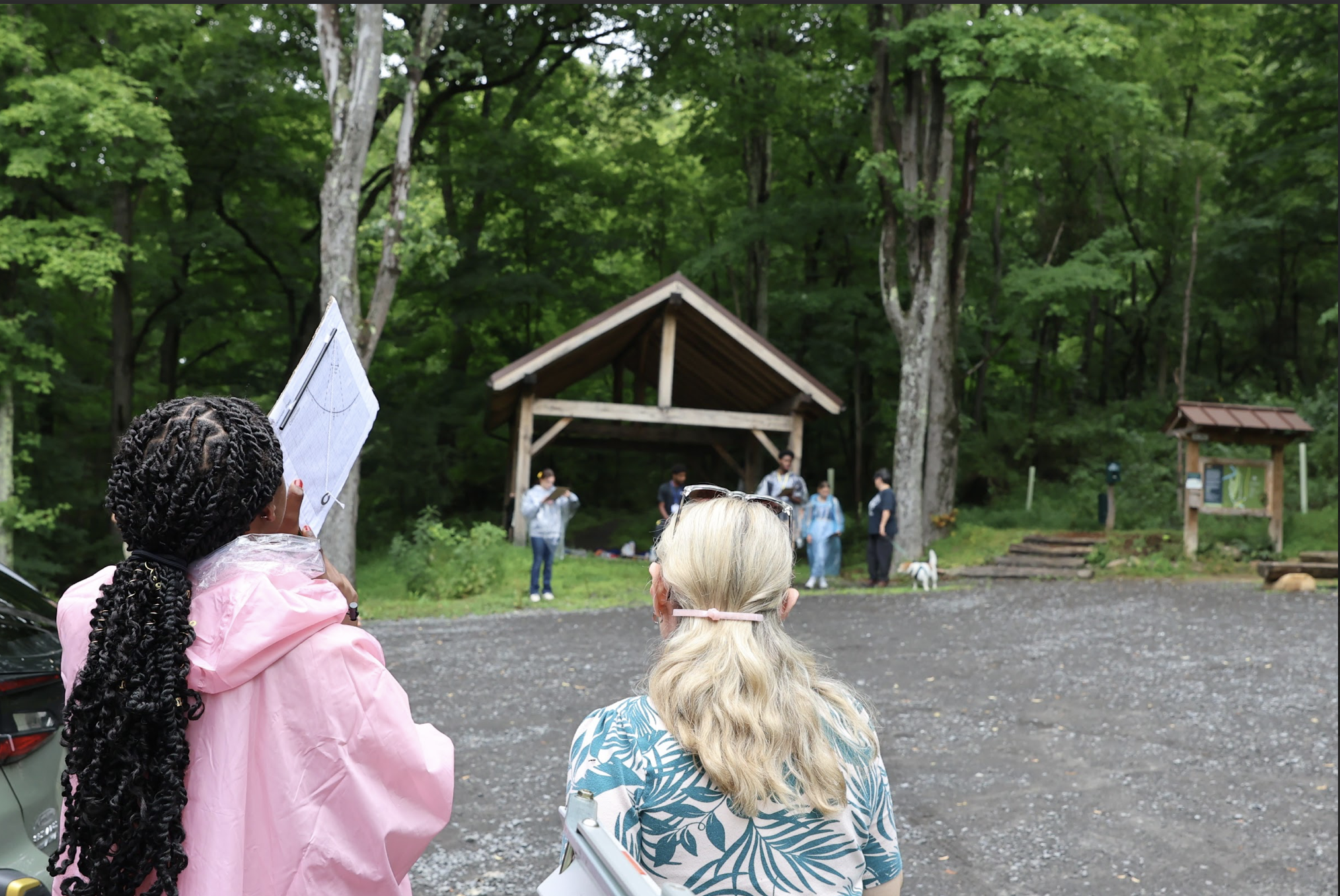 one student in the foreground uses a clinometer to measure the height of a tree in the background