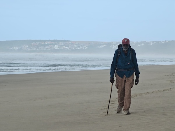 Dr. John Francis walks along a beach in South Africa wearing a backpack and walking with a stick