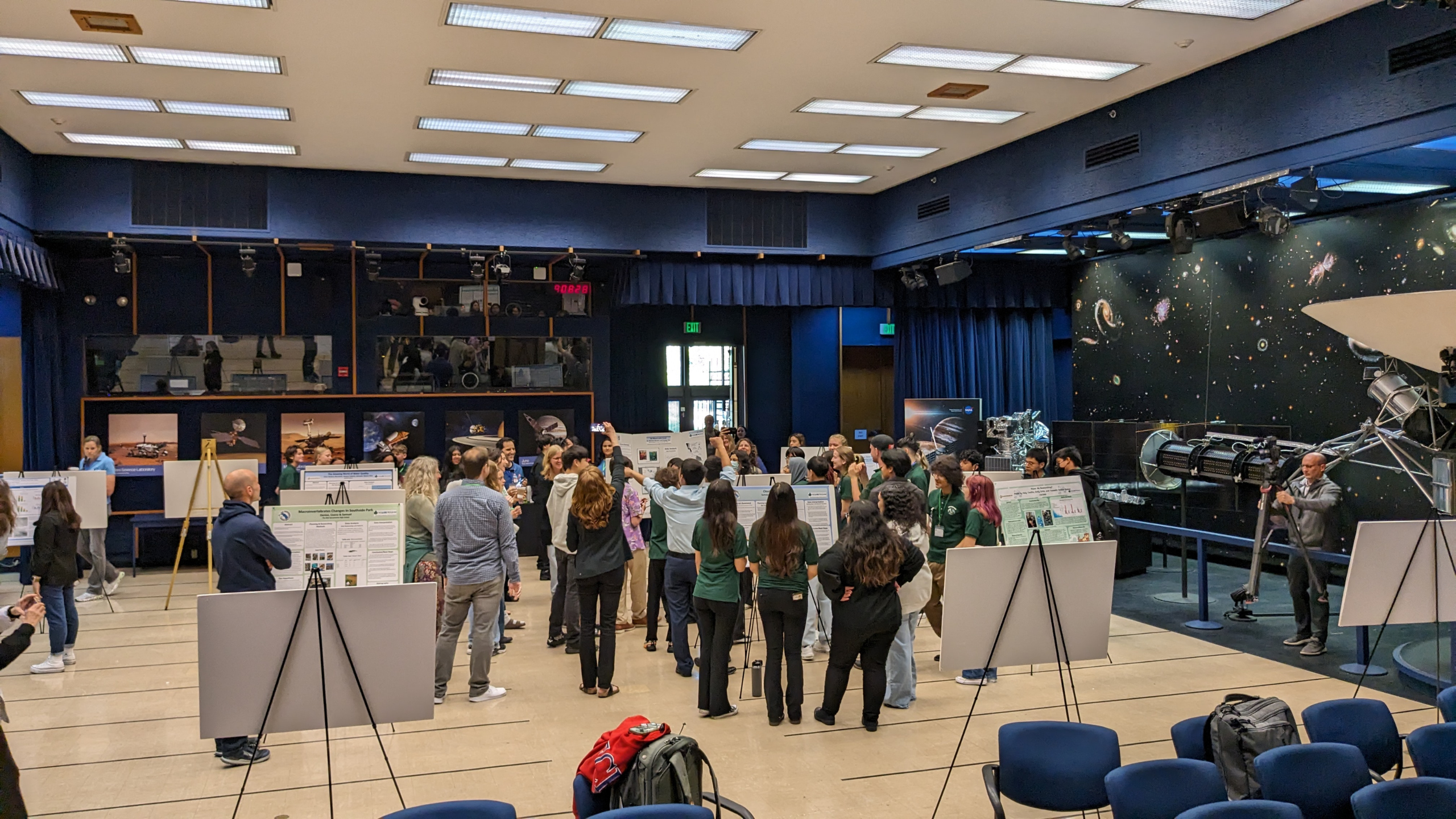 Students huddled in the middle of a large room, surrounded by their research posters on easels, as they participated in an ice breaker on day 1 at the Pacific Region Student Research Symposium