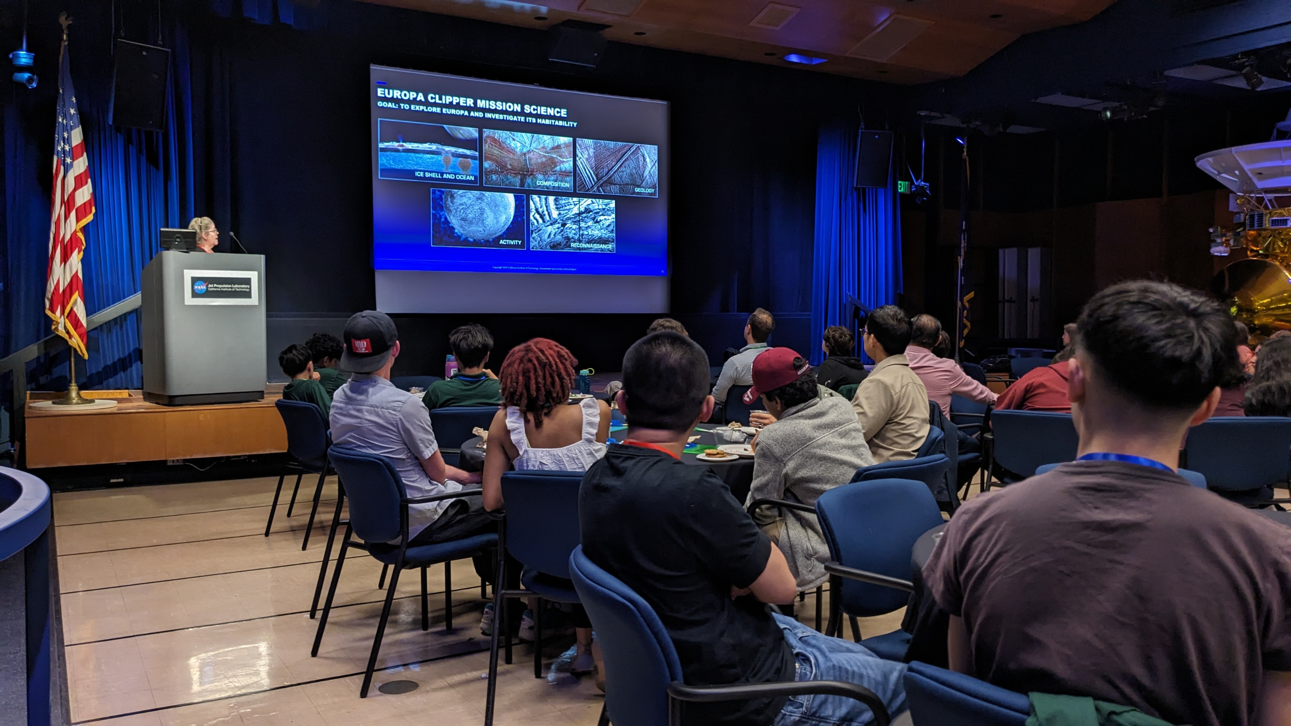 Ocean McIntyre stands at a podium to give a keynote speech about the Europa Clipper satellite mission while educators and students look at a screen with slides