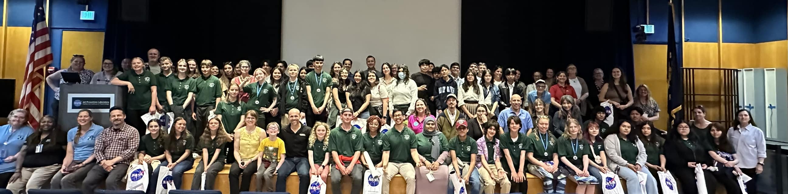 Approximately 100 GLOBE students, educators, Partners and chaperones sit or stand on a stage at the 2024 Pacific Region Student Research Symposium