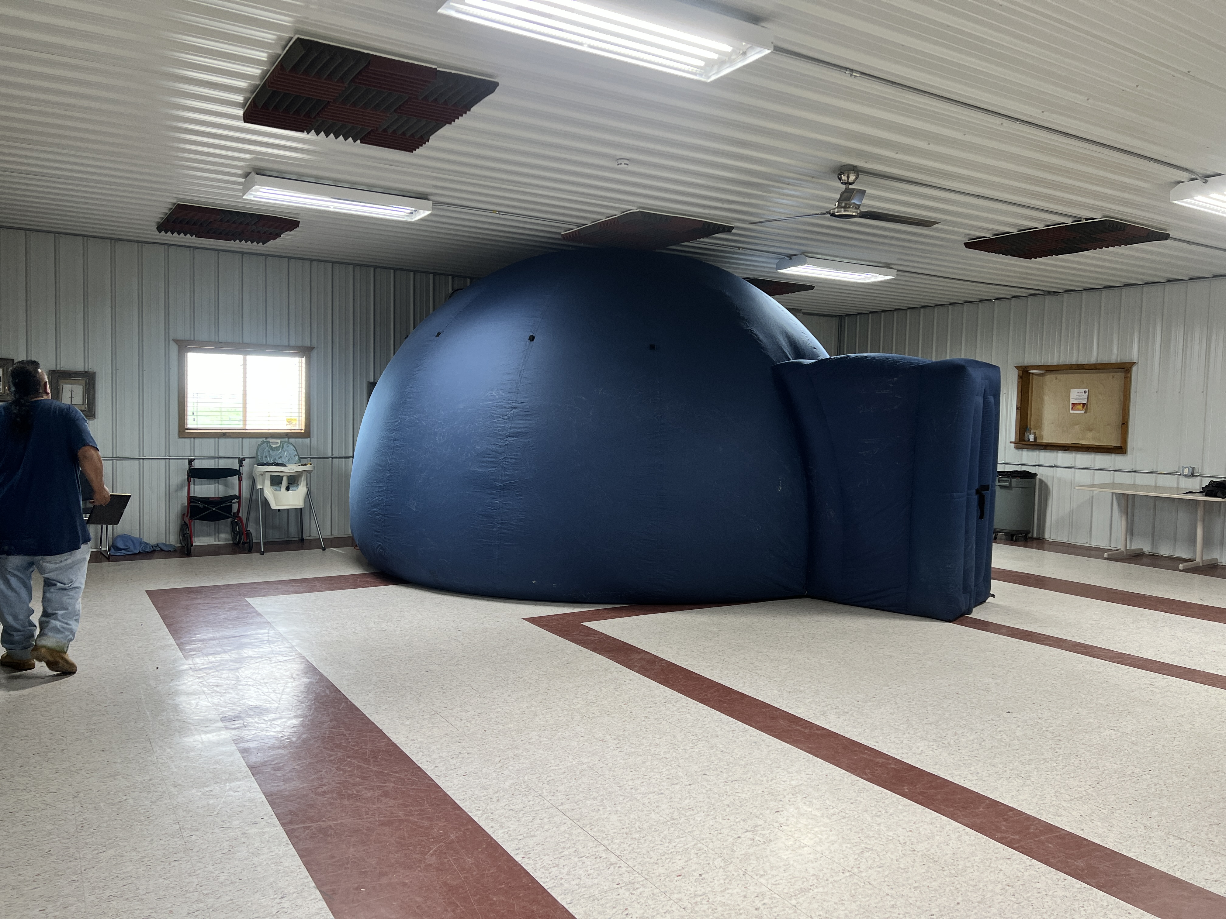 A StarLab indoor planetarium exhibit is in the middle of a room and its roof touches the ceiling. It has a small entryway that leads to a rounded viewing area. It appears to be about 9 feet tall and 9 to 10 feet wide.