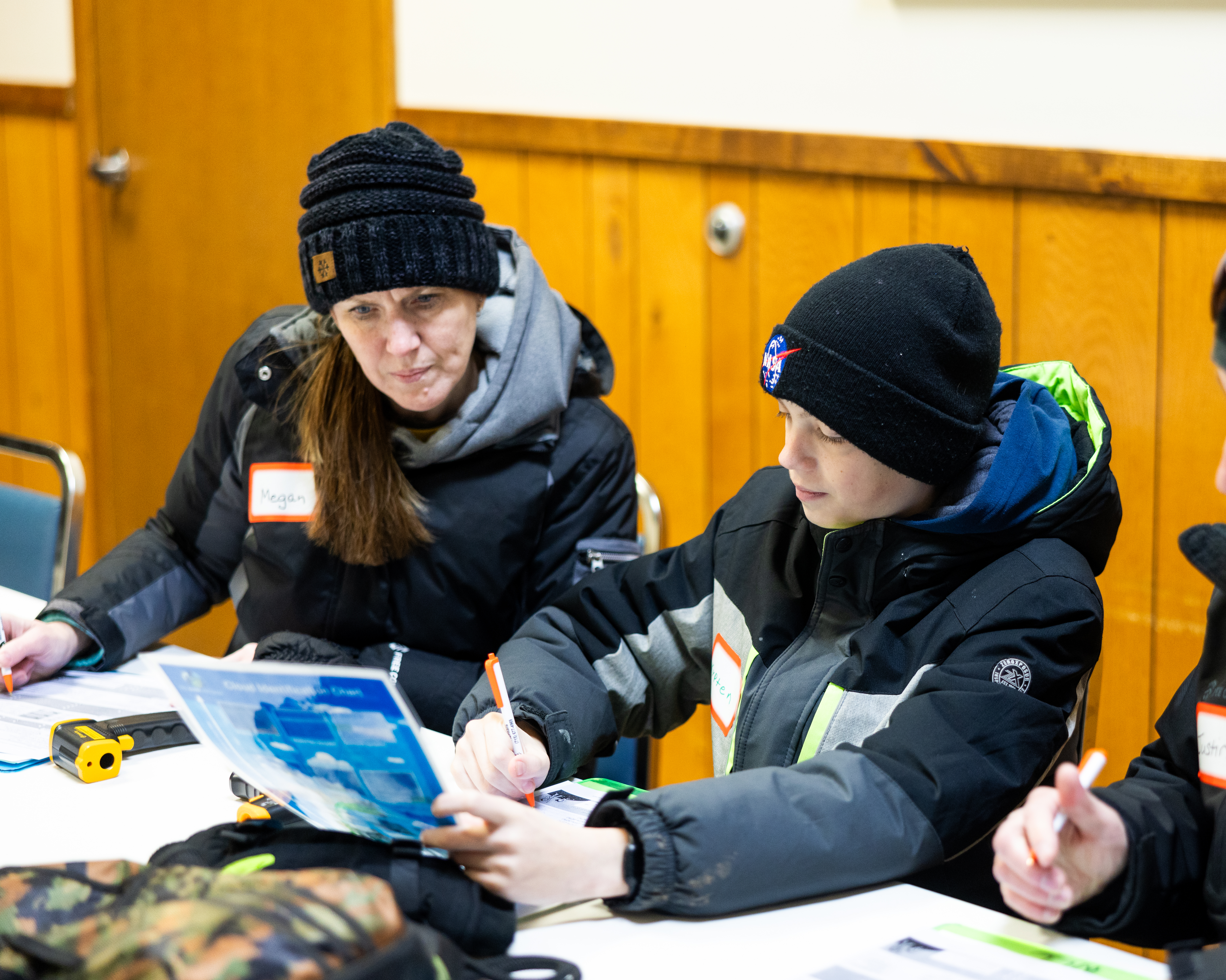 A woman dressed in a coat and hat sits at a table next to a boy also dressed in a coat and hat. The boy is holding a cloud chart and writing on a paper. The woman is looking at the chart and is also writing on a paper in front of her. An infrared thermometer is on the table between them. 