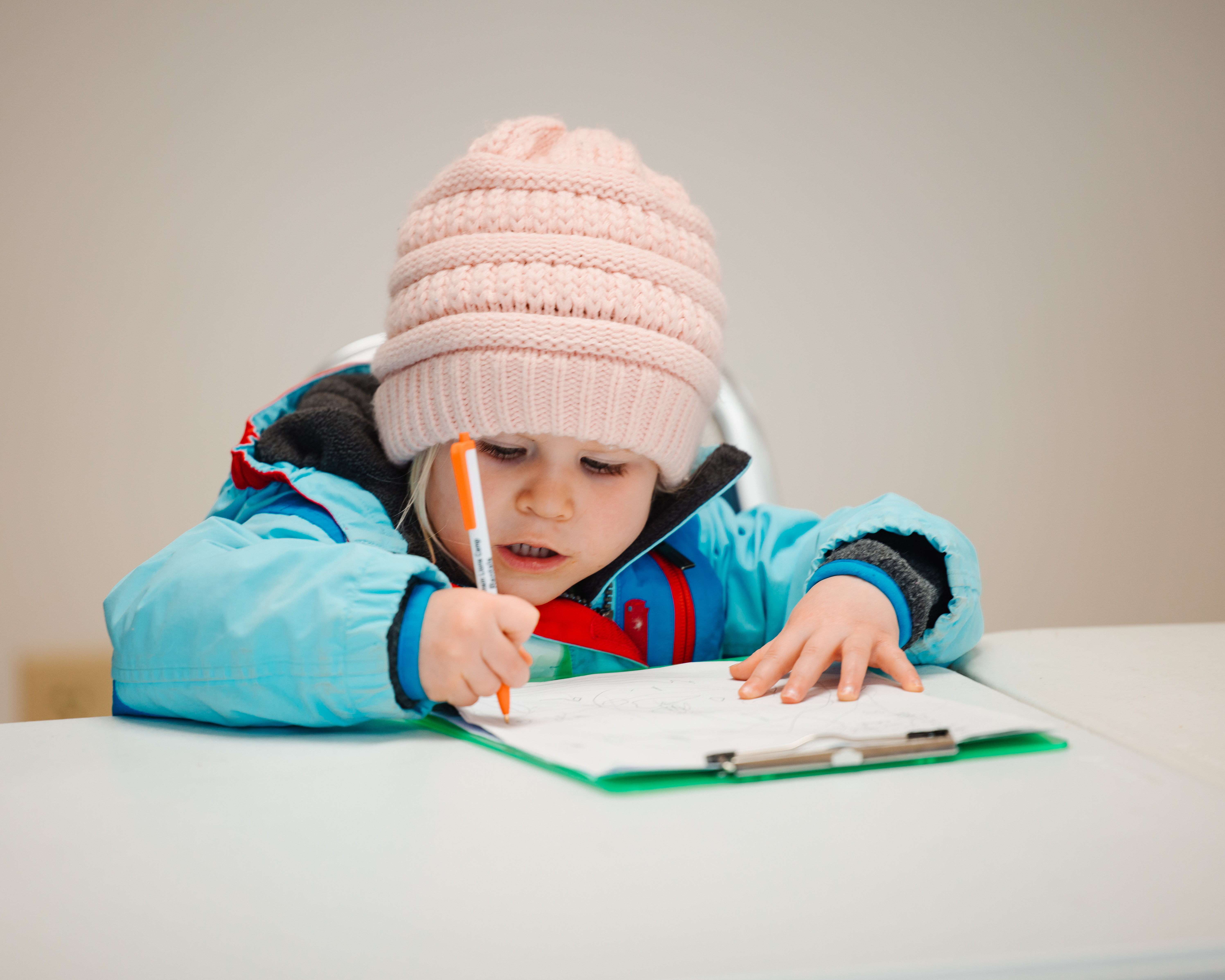 A preschool-aged girl wearing a coat and a hat sits at a table and draws on a data sheet with a pen. 