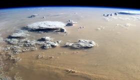 A photograph of Earth from the International Space Station shows a large dust storm coloring the skies brown. Towering thunderstorm clouds rise above the dust. The Earth's blue limb is at the top of the photo.