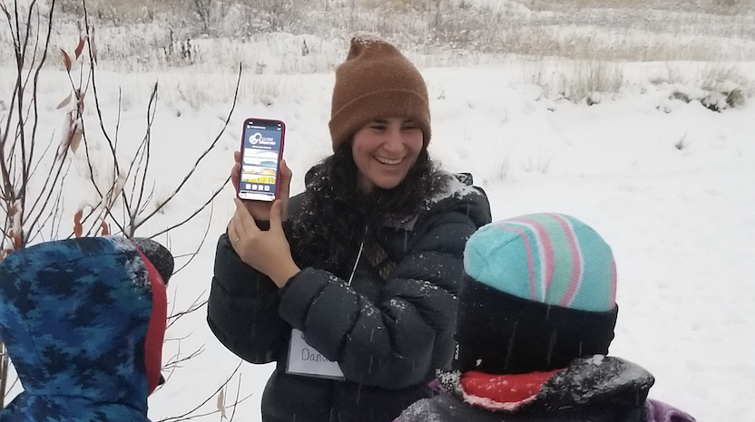A photo of a woman in a winter coat and hat showing a phone screen to two children. The screen shows the GLOBE Observer app. All are standing outside on a snow-covered landscape.