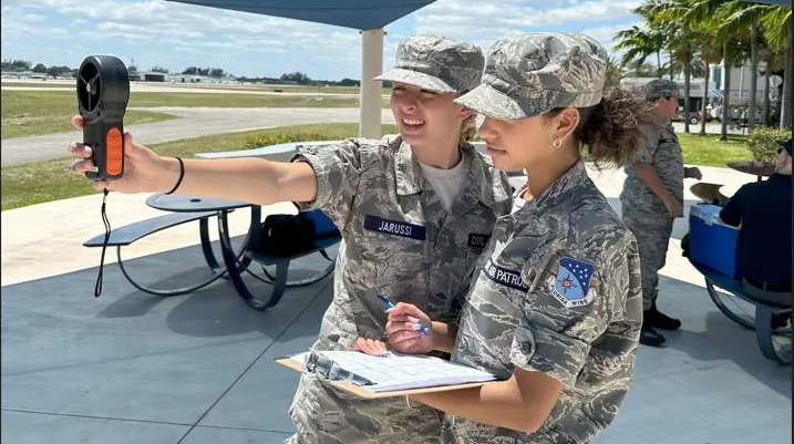 Two Civil Air Patrol cadets stand collecting data with an anemometer and a clipboard.