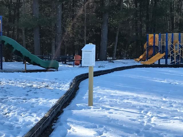 Picture of a weather shelter in an open area with snow. It is near a playground with a tree-line in the background. 
