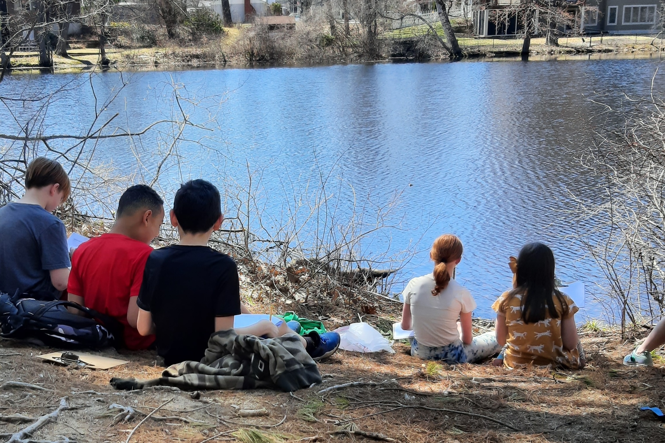 Five kids write on clipboards at the edge of a river in the early spring.