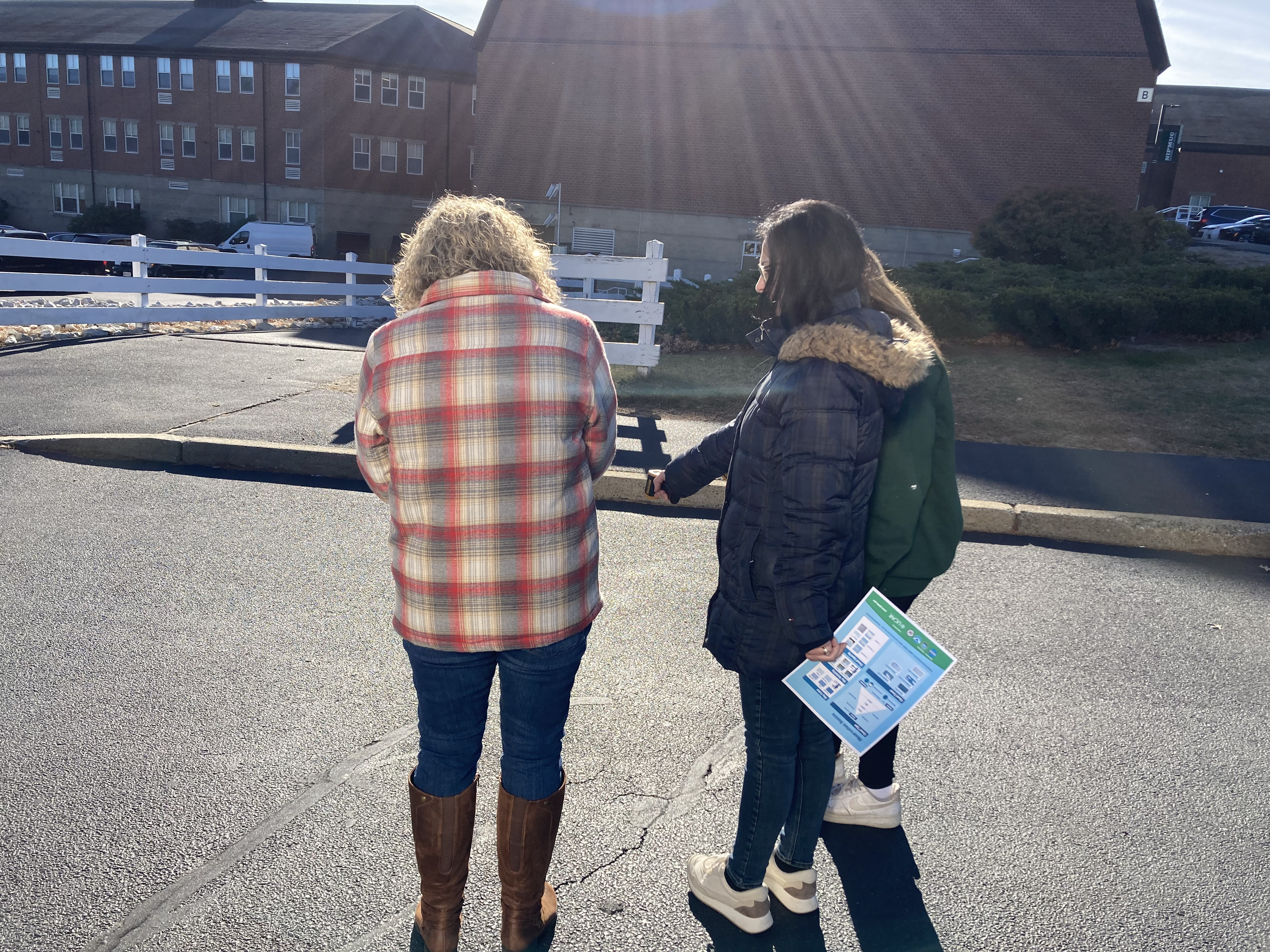 three people stand on a paved parking lot near brick buildings; the sun is shining as they practice taking surface temperature measurements with an infrared thermometer